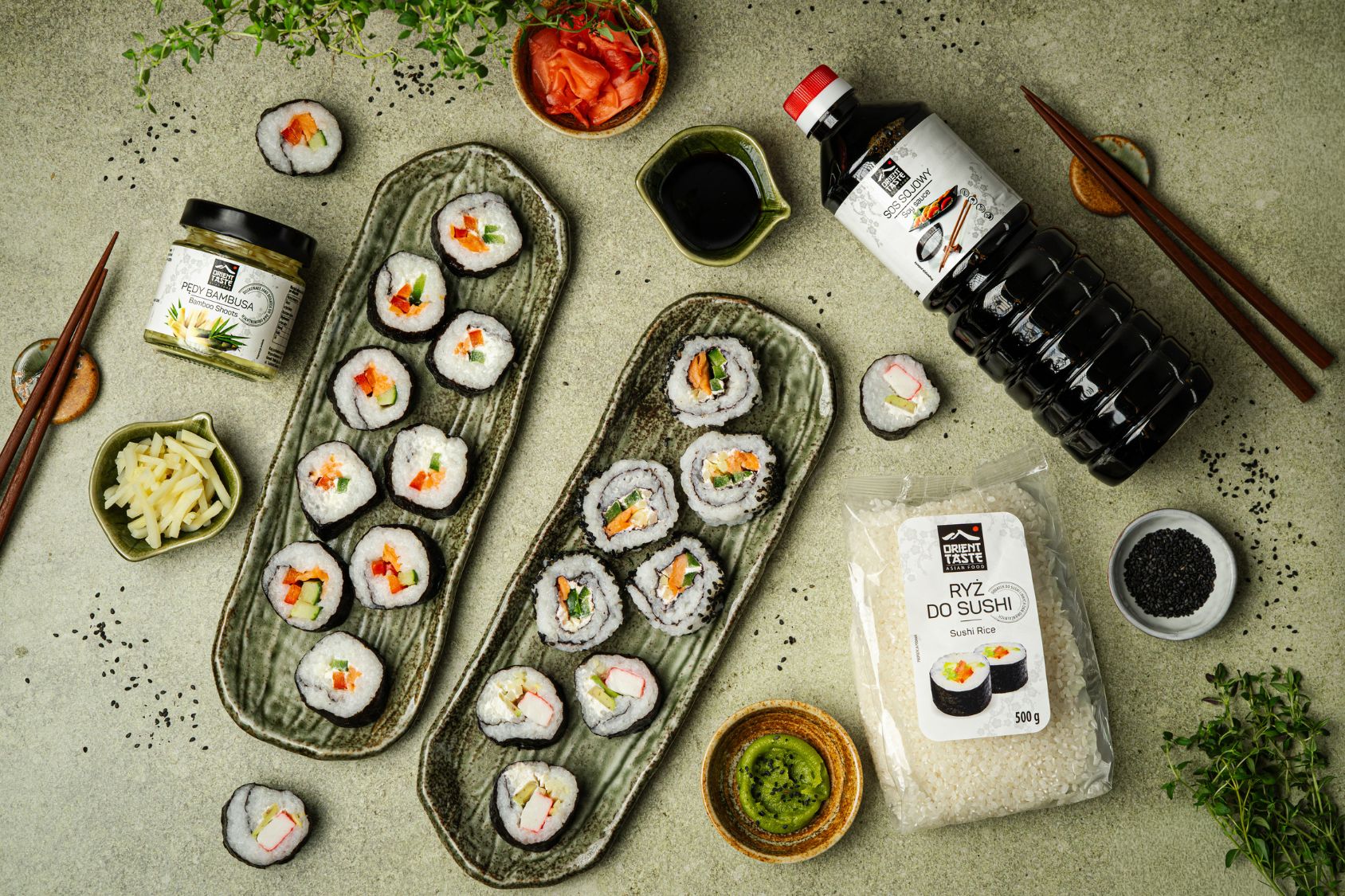 Gotowe sushi, leży na stole na dekoracyjnych podłużnych talerzach. Obok na stole leżą produkty od Orient Taste, sos sojowy, ryż do sushi, pędy bambusa.